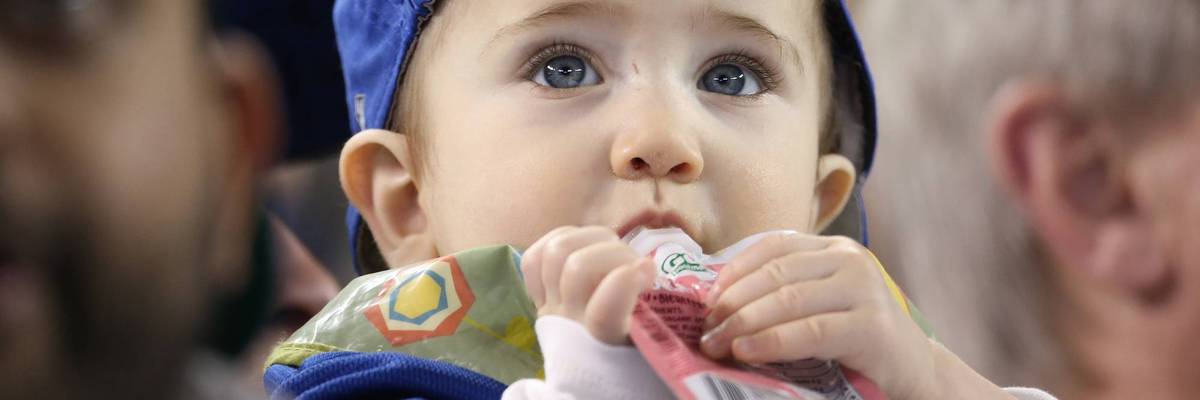 A baby eats from a plastic pouch while watching baseball.