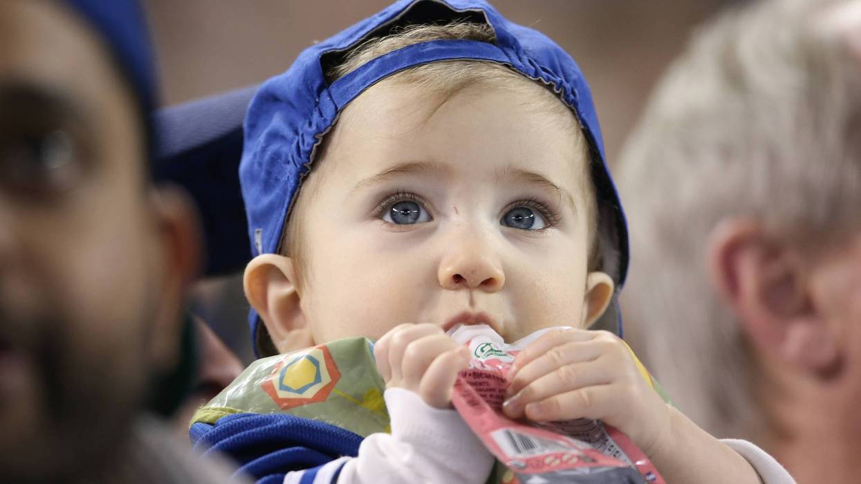 A baby eats from a plastic pouch while watching baseball.
