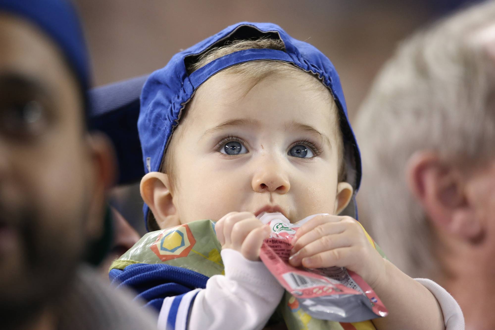 A baby eats from a plastic pouch while watching baseball.