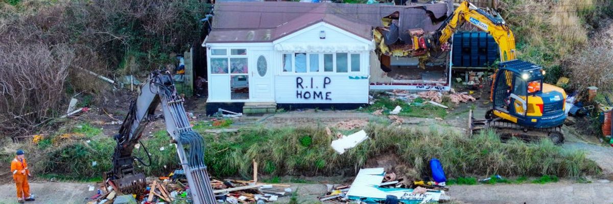 A beach house adorned with "RIP HOME" is demolished by workers before it falls down the sea cliff