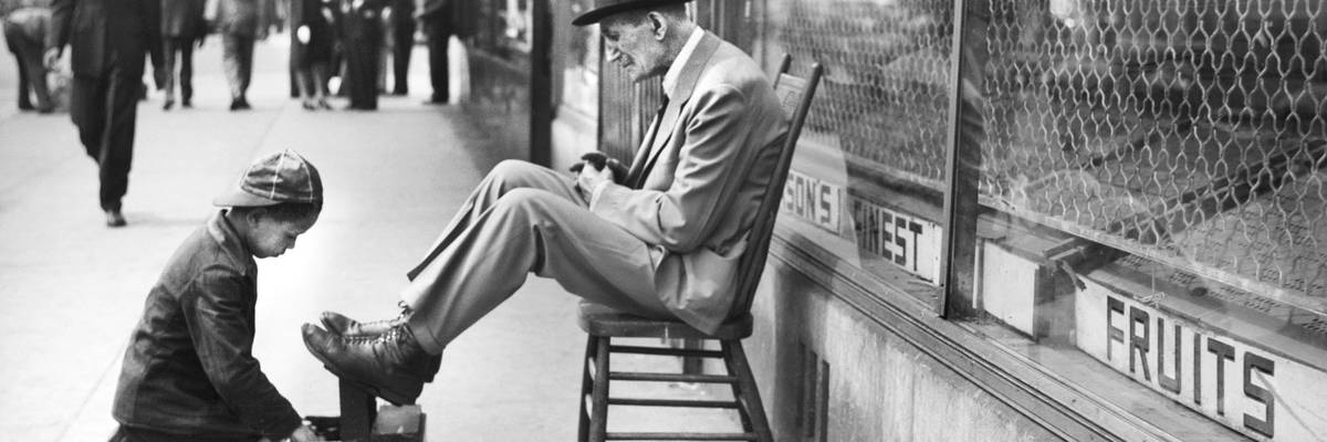 A black-and-white image of a boy kneeling before a man to shine his shoes.