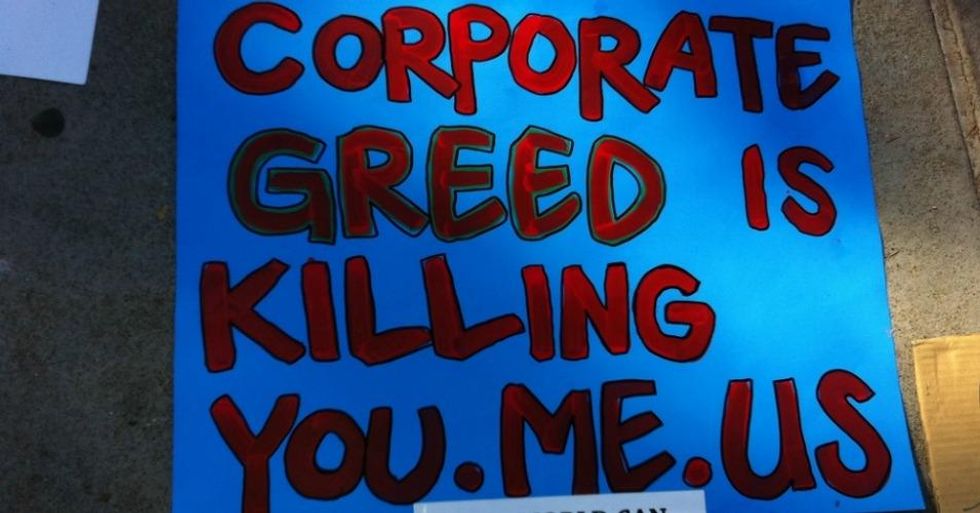 A blue sign reading "Corporate greed is killing You. Me. Us."