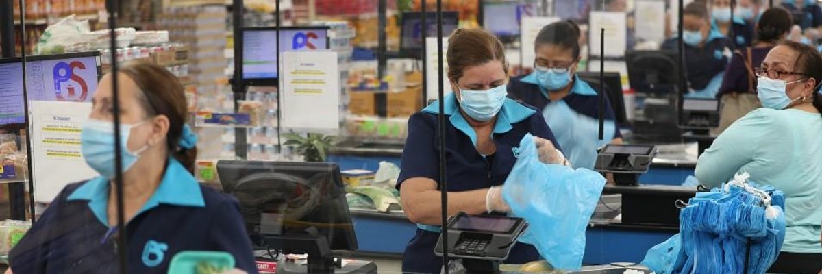 A cashier stands behind a partial protective plastic screen and wears a mask and gloves at the Presidente Supermarket on April 13, 2020 in Miami.