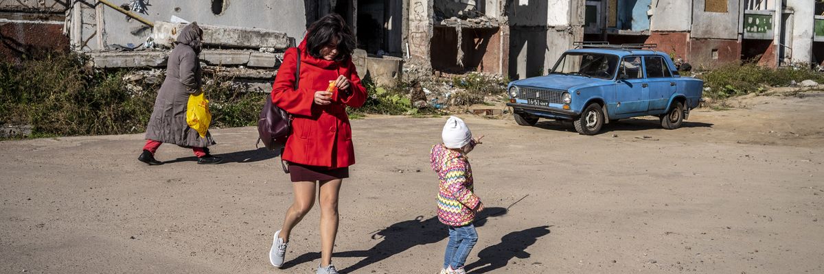 A child walking with a woman points at an old blue car seen amid severely damaged buildings