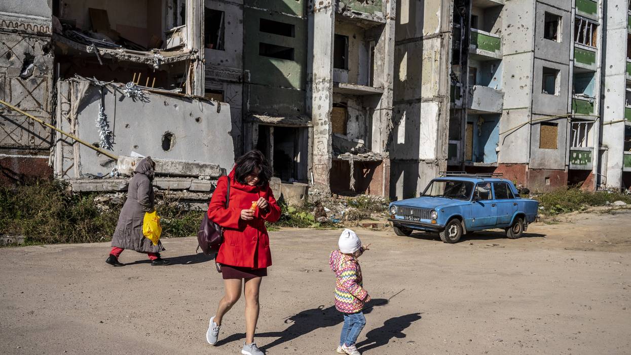 A child walking with a woman points at an old blue car seen amid severely damaged buildings