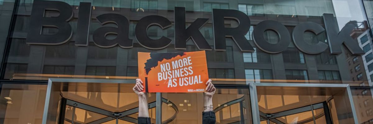 A climate activist holds a sign saying, "No more business as usual," in front of the BlackRock logo.