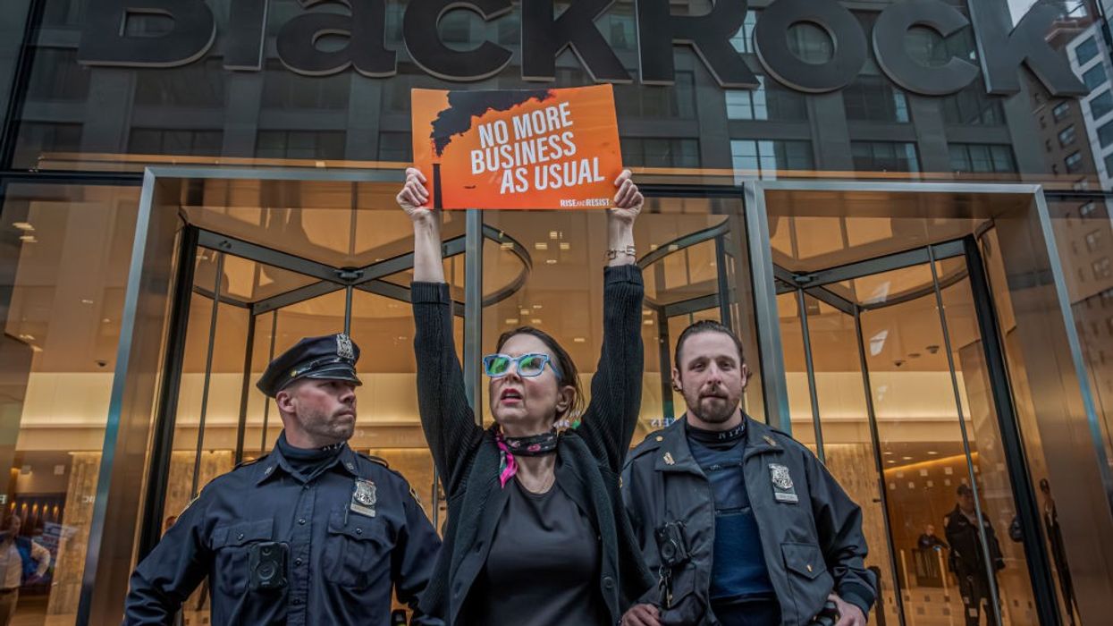 A climate activist holds a sign saying, "No more business as usual," in front of the BlackRock logo.