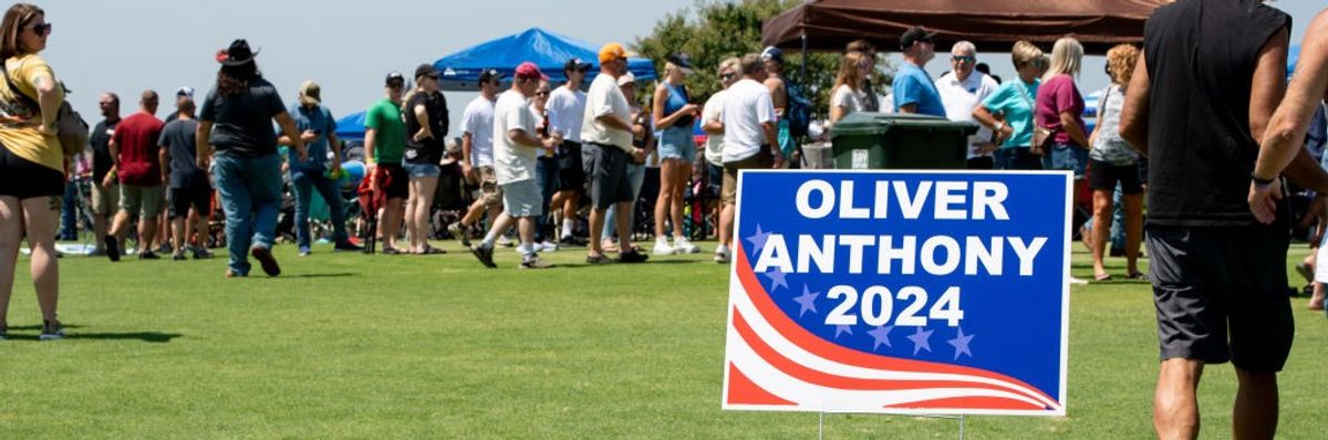 A crowd gathers on a lawn behind a sign reading, "Oliver Anthony 2024."