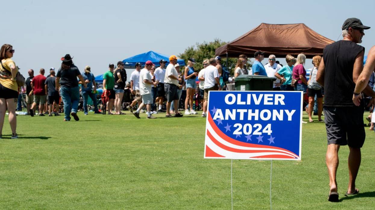 A crowd gathers on a lawn behind a sign reading, "Oliver Anthony 2024."