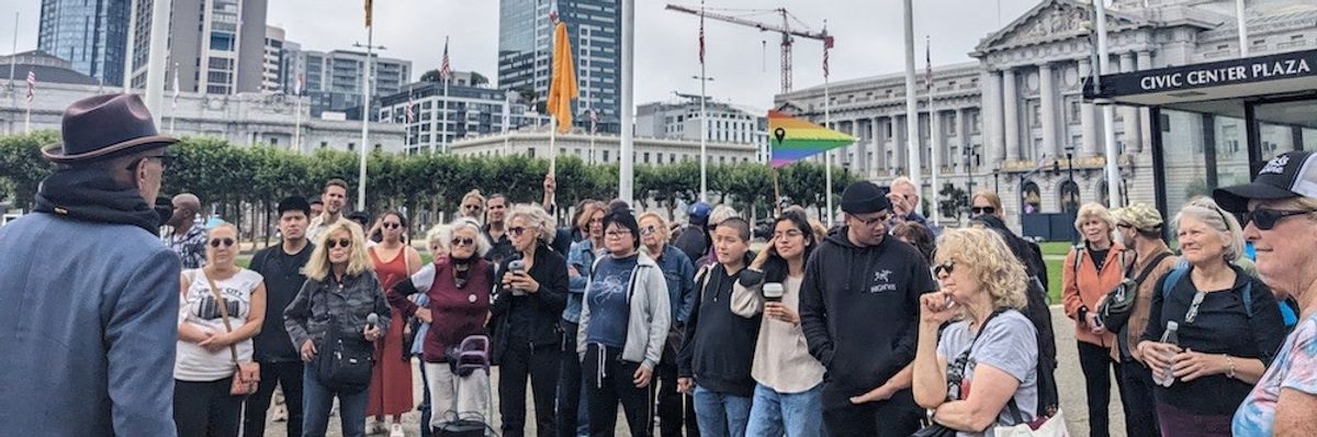 A crowd listens to a man in a fedora and scarf speak. 