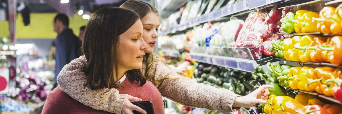 A daughter reaches for bell peppers in a store while piggy-back riding on her mother's shoulders. 