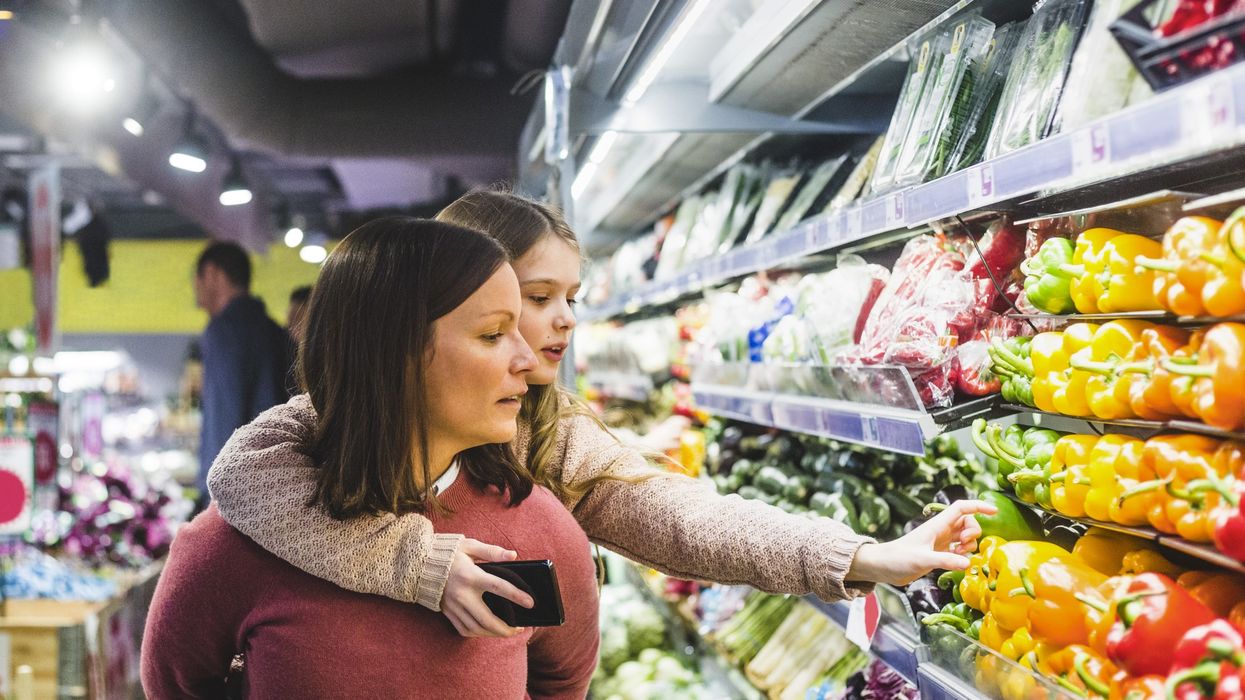 A daughter reaches for bell peppers in a store while piggy-back riding on her mother's shoulders.