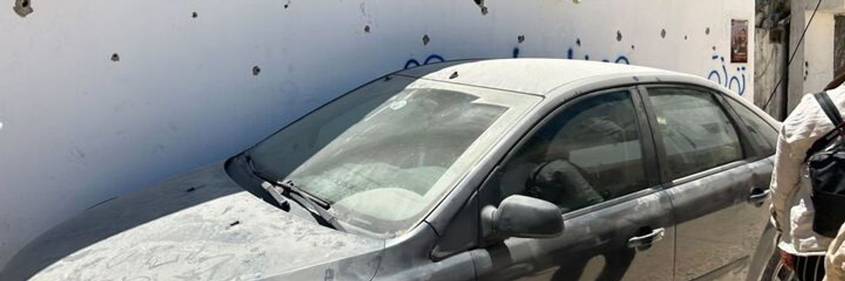 A dusty car against a white wall with bullet holes.