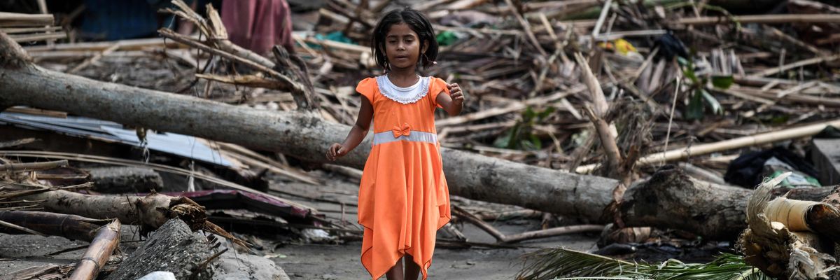 A girl in an orange dress stands amidst rubble.