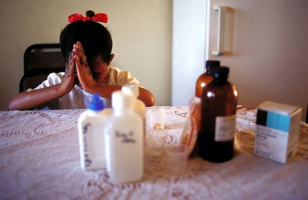 A girl with sores on her face and a red bow in her hair bows her head in prayer; pill bottles are seen in the foreground