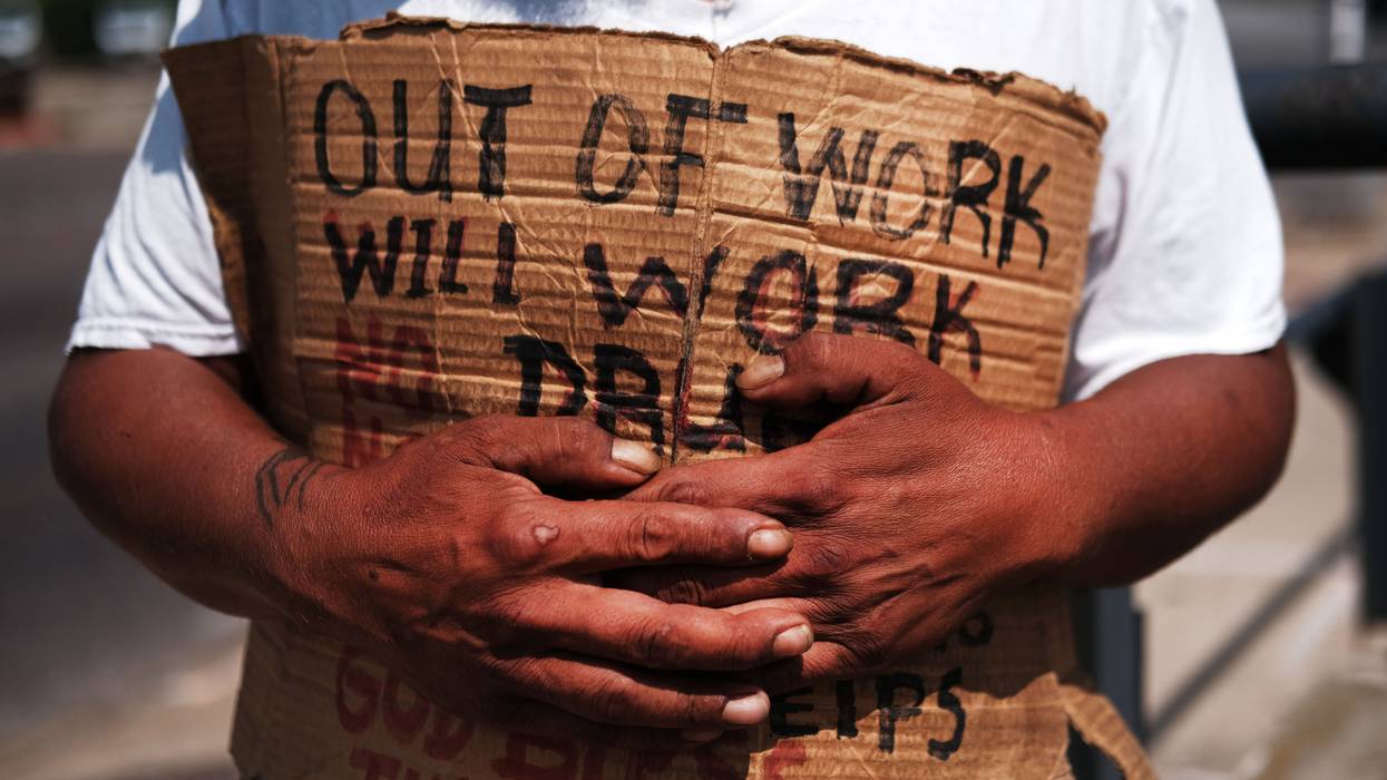 A homeless man holds a cardboard sign reading, "Out of work. Will work."