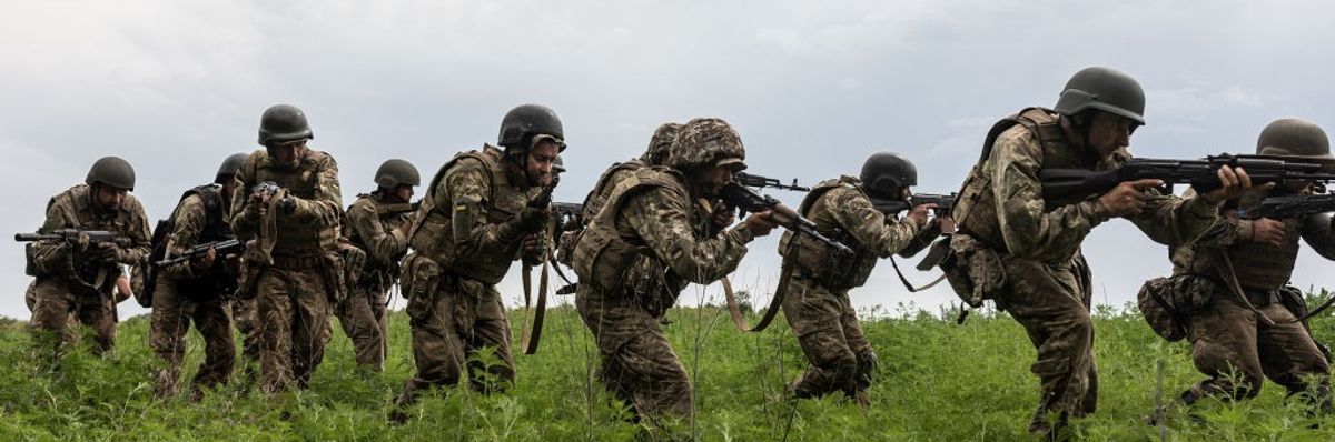 A line of Ukrainian soldiers crouches in line in a field with guns. 