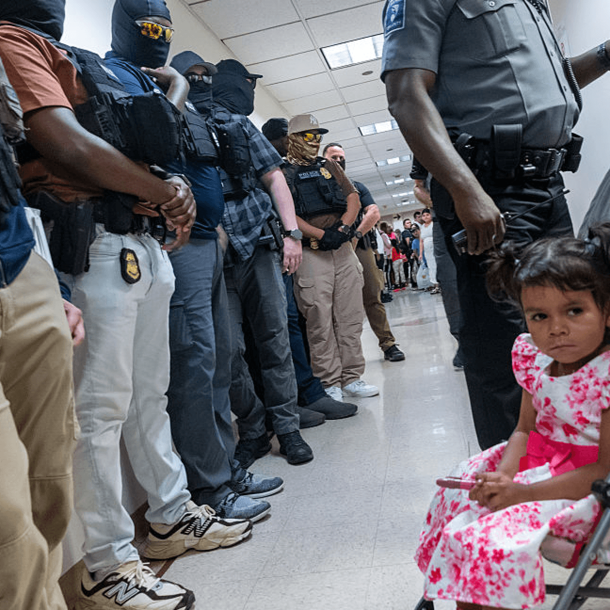 A little girl sits in a stroller in front of federal immigration enforcers at a courthouse