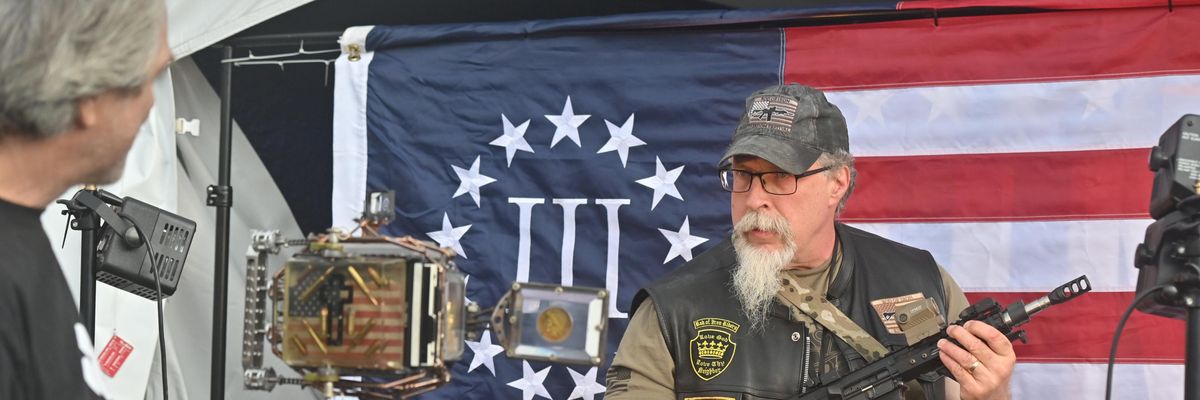 A man holding an automatic rifle poses in front of a 3% flag, representing a far-right Christian fundamentalist worldview
