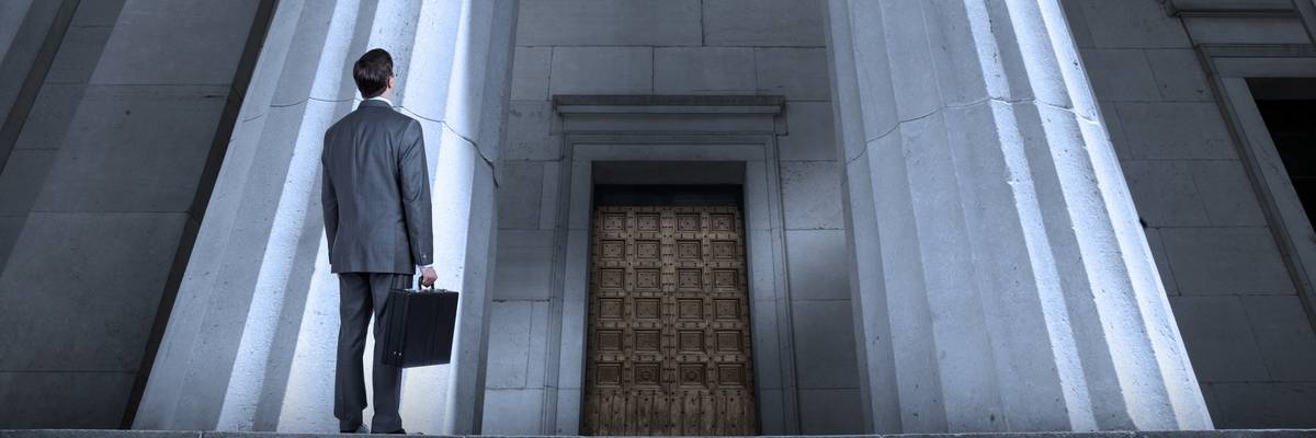 A man in a suit with a briefcase looks up at a courthouse. 