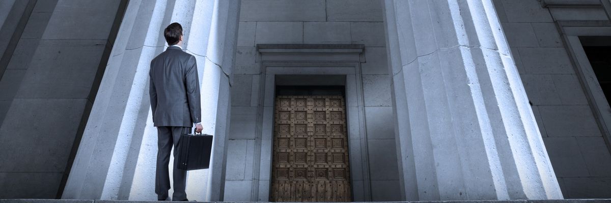 A man in a suit with a briefcase looks up at a courthouse. 