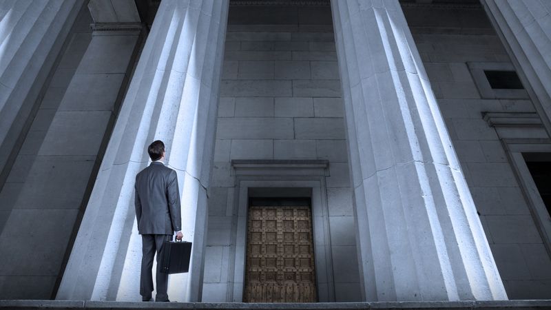 A man in a suit with a briefcase looks up at a courthouse. 