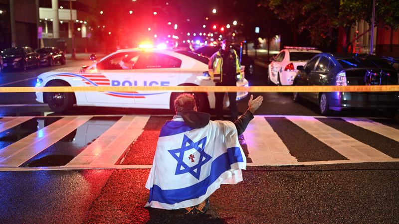 A man in an Israeli flag stands in front of a police line and car.