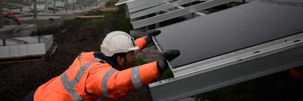 A man in an orange jacket installs a solar panel on a roof. 