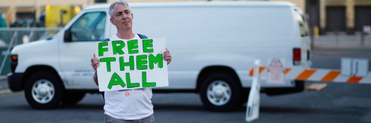 A man stands in front of a white van with a white sign reading, "Free them all," in green.