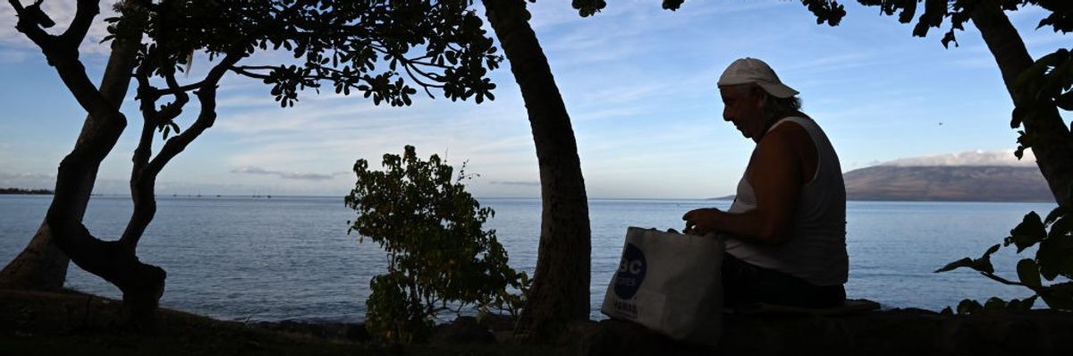 A man stands with his belongings in a paper bag against the ocean. 
