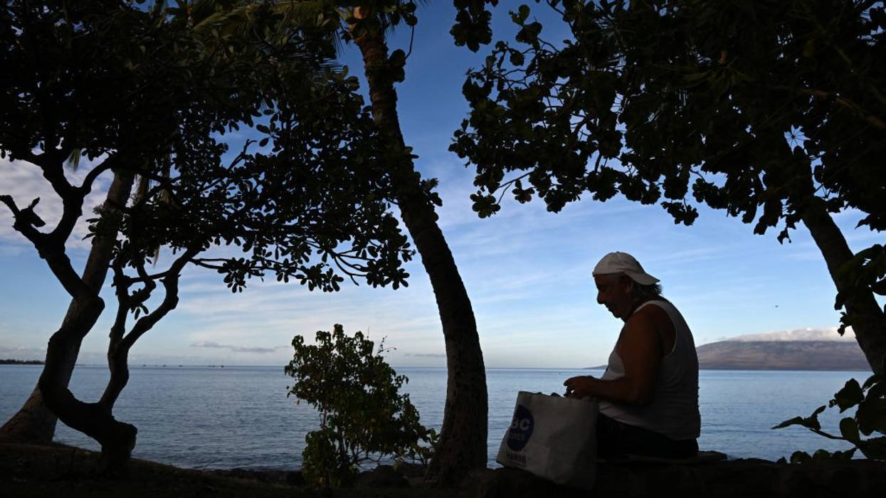 A man stands with his belongings in a paper bag against the ocean.