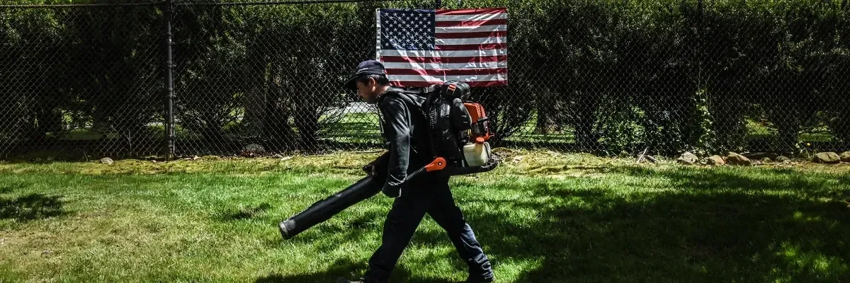A person holding a leaf blower walks past an American fag.