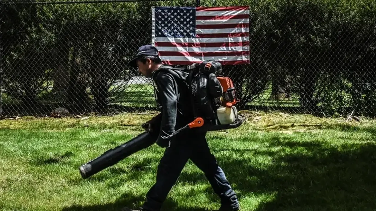 A person holding a leaf blower walks past an American fag.