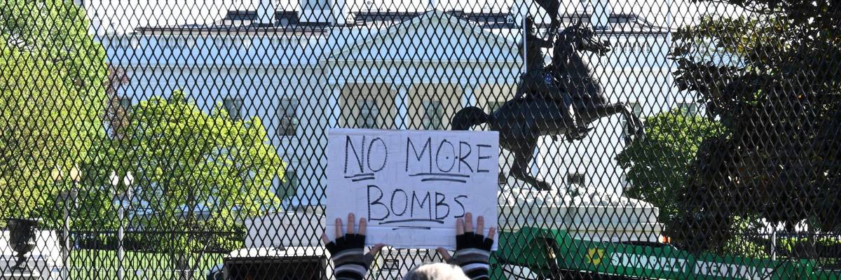 A person holds a sign up to a fence in front of the White House reading, "No more bombs."