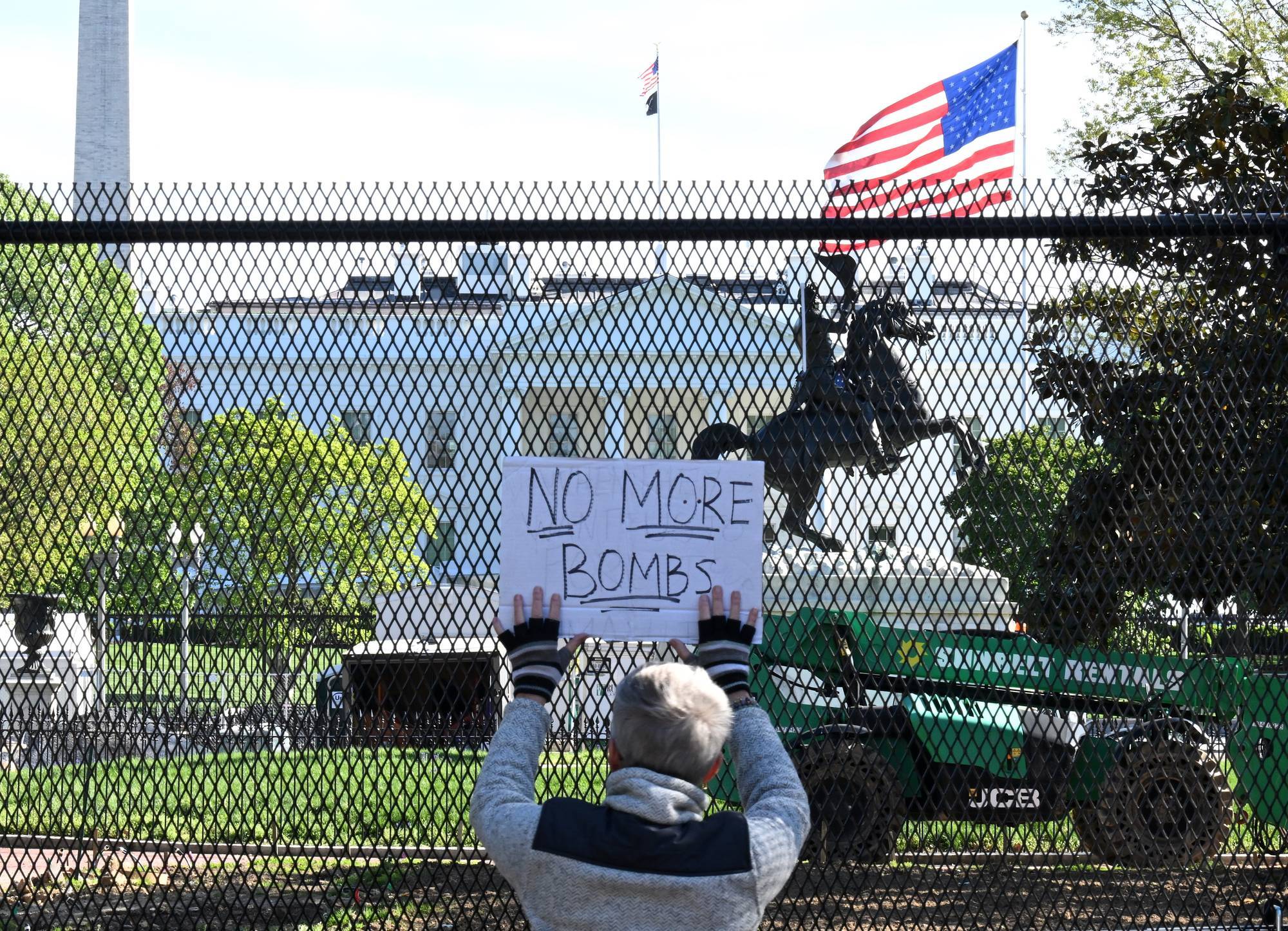 A person holds a sign up to a fence in front of the White House reading, "No more bombs."