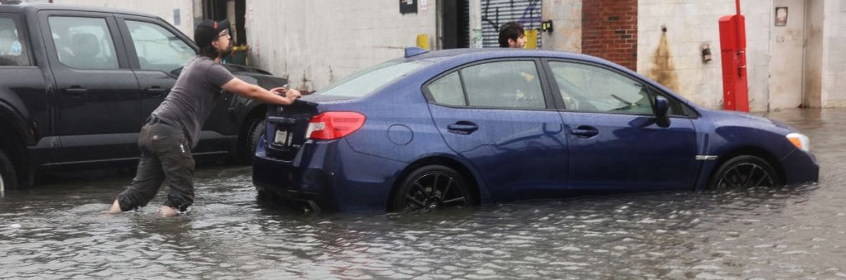 A person in a gray t-shirt pushes a blue car through a flooded street.