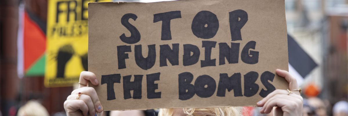 A protester holds a cardboard sign reading, "Stop funding the bombs."