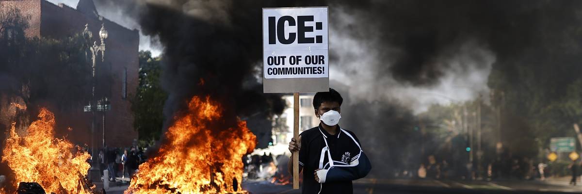 A protester holds a sign reading: "ICE: Out Of Our Communities" as burning Waymo cars line the street
