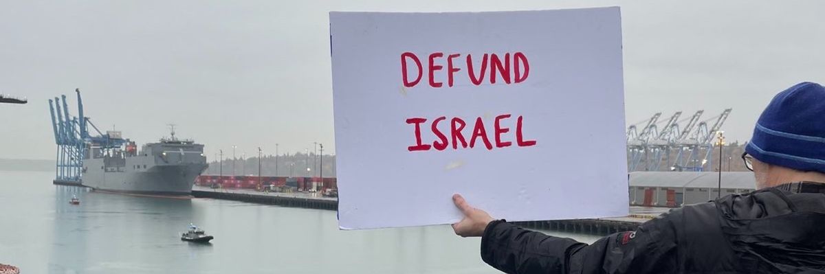 A protester holds out a sign reading, "Defund Israel" as the MV Cape Orlando is shown in the distance.