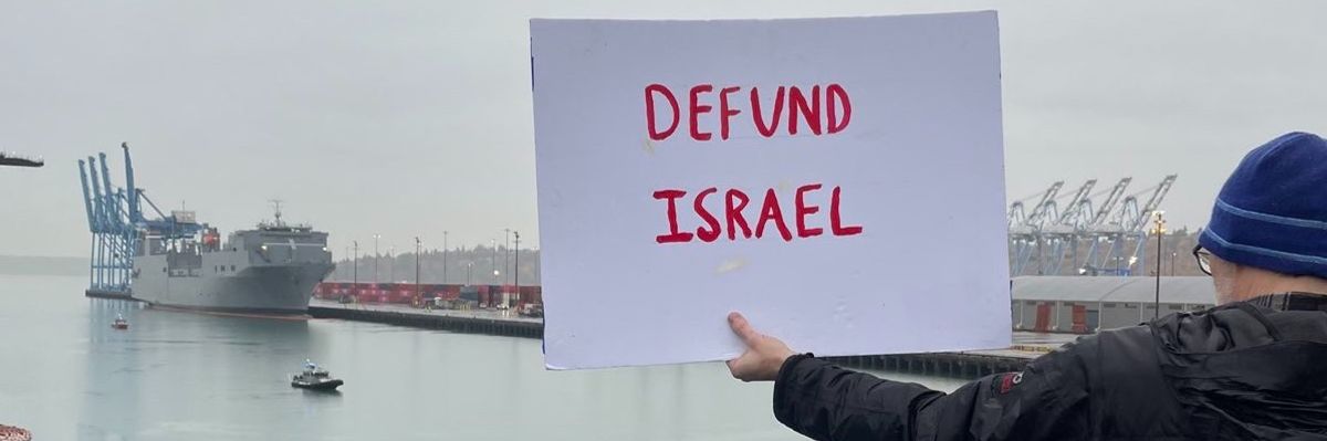 A protester holds out a sign reading, "Defund Israel" as the MV Cape Orlando is shown in the distance. 