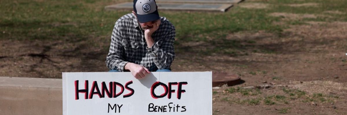 A protester sits with a sign reading, "Hands off my benefits, VA, Social Security, Medicare,"