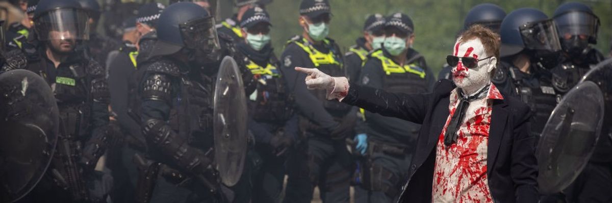 A protesters in a suit smeared with fake blood points toward Victoria police in riot gear