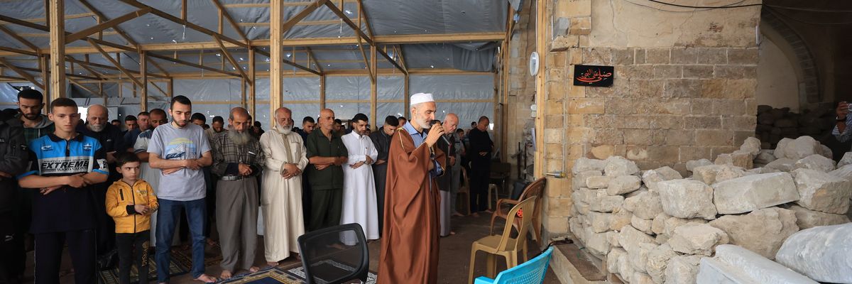 a row of men, with heads bent in prayer, stand behind a man with a microphone.