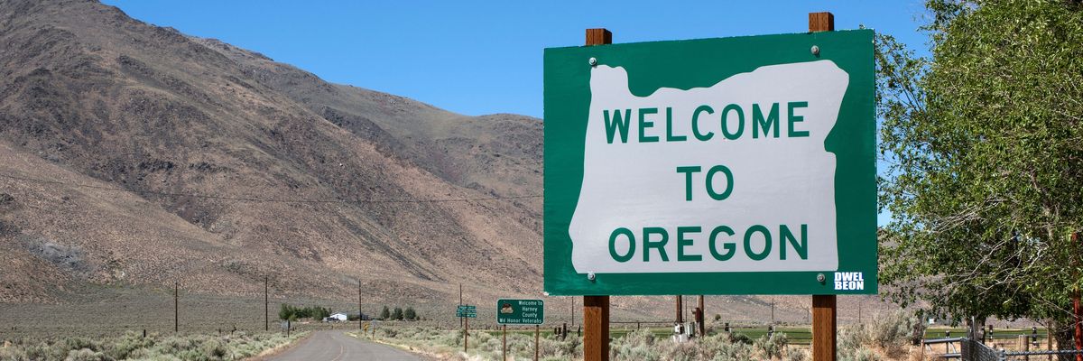 A sign reads "Welcome to Oregon" along a two-land country road with a mountainout backdrop.
