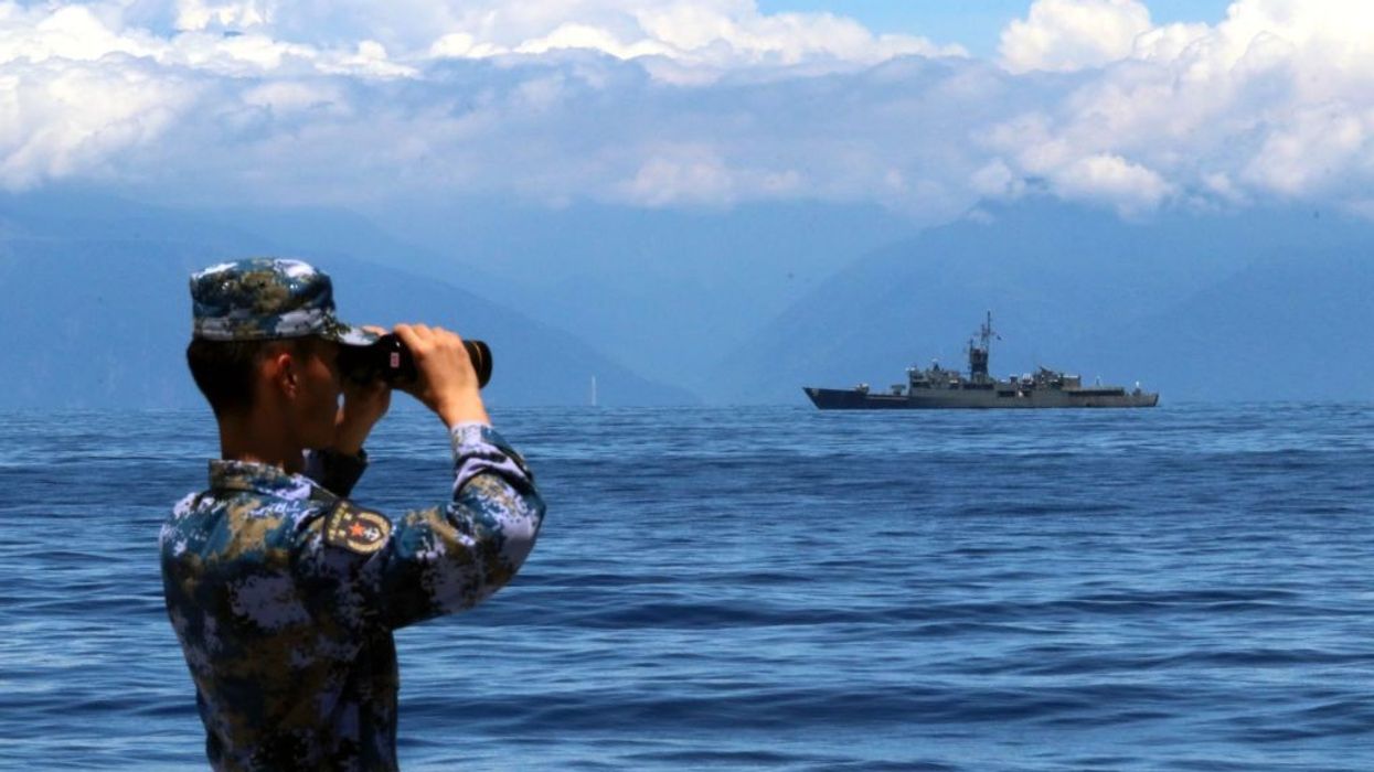A soldier looks through binoculars at a naval ship on the water.