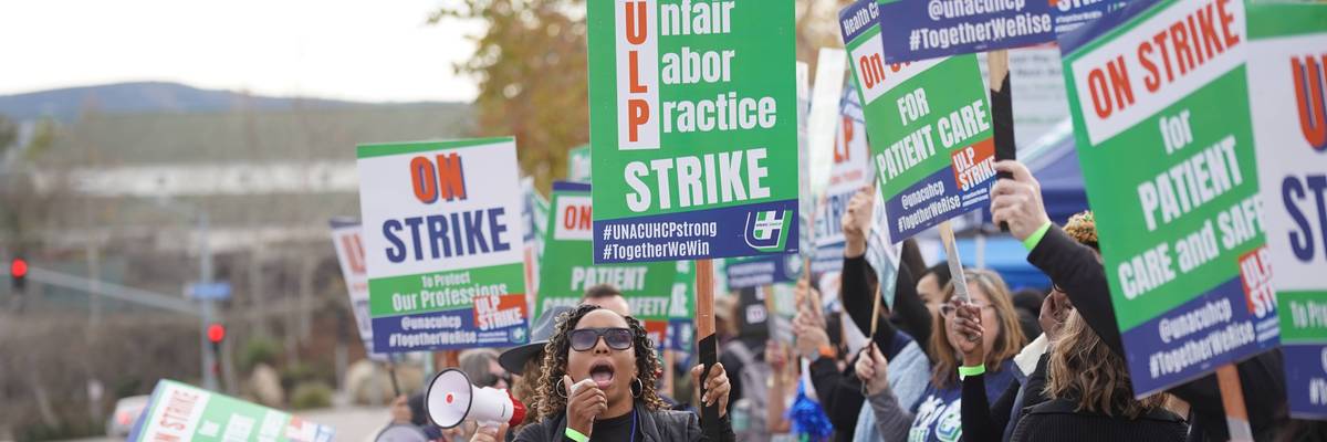 A striking Kaiser nurse speaks into a megaphone while holding a sign reading, "Unfair Labor Practice Strike"