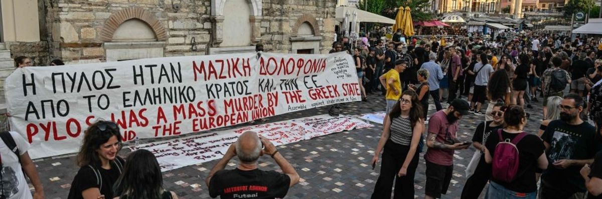 A white banner with red and black writing displayed in a square in Athens with a crowd gathered around it.