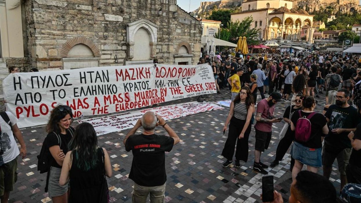 A white banner with red and black writing displayed in a square in Athens with a crowd gathered around it.