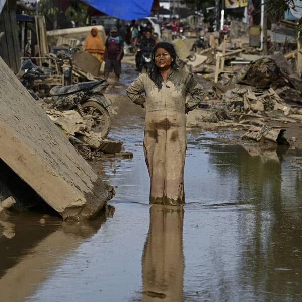A woman covered in mud stands on a street surrounded by debris