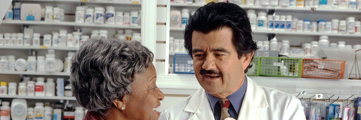 A woman in red speaks to a man in a white coat who holds a yellow pill bottle.